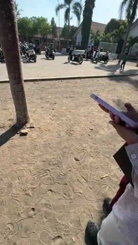 The video begins with a close-up view of dusty ground covered in shoe prints. The camera then pans up to reveal two young women standing in what appears to be an outdoor area, possibly a schoolyard or parking lot. Both women are wearing hijabs. The woman in the foreground, closer to the camera, is wearing glasses and a checkered shirt. She is smiling and adjusting her hijab. The second woman, slightly behind and to the left, is wearing a pink hijab and glasses, and is looking down at her phone. In the background, there is a large number of parked motorcycles, several trees, and buildings with tiled roofs. A soccer goal is also visible in the middle ground. The sky is clear and blue, indicating a sunny day. The overall impression is of a casual outdoor setting on a bright day.