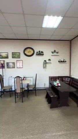 The video pans from the right to the left, showing the interior of a small establishment. In the foreground, there is a dark wooden booth-style table with two matching benches. To the left of this table, there is a small square table with two chairs around it. The walls are decorated with framed pictures and a large clock. Further to the left, there is a counter with a refrigerated display case filled with food items. Behind the counter, there are shelves displaying various products. A tall, dark pillar stands in the middle of the room. On the left side of the room, there is a refrigerator filled with bottled drinks and a sink area. The floor is covered with a textured material, and the ceiling has recessed lighting. The establishment appears to be a cafe or a small restaurant.