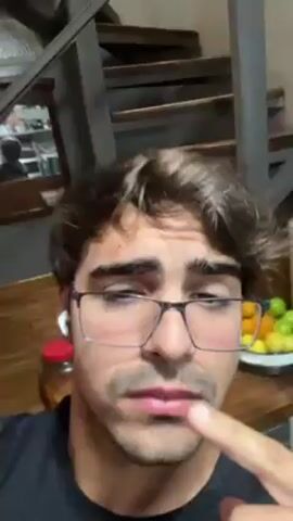 A man wearing glasses and earbuds looks at the camera. He touches the bridge of his glasses with his finger, then touches his jaw. A wooden staircase is in the background, and a plate of fruit is on the table. He is wearing a black t-shirt.