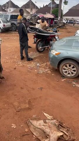 The video shows an outdoor scene on a dirt lot with several people, vehicles, and structures. Initially, three men are grouped around a parked motorcycle, seemingly adjusting a large, dark object on it. Nearby are a silver car and other vehicles. The camera then pans down to show a dirty, tattered sack on the ground amidst scattered trash. Following that, the camera focuses on a makeshift structure constructed from corrugated metal and wood, with a man walking behind it. In the background, a bustling market scene is visible with more people, cars, and vendor umbrellas. The ground throughout the scene is red dirt with scattered debris.