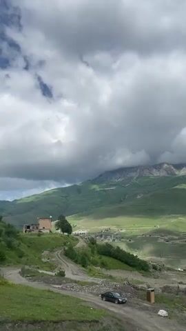 This video shows a scenic view of a mountainous landscape in Verkhny Fiagdon, Russia. The scene is dominated by lush green hills and majestic mountains, with a cloudy sky overhead. In the foreground, a dirt road winds its way down a hillside, with a dark-colored car parked on it. A few buildings are visible on the slopes, including a small brick house. In the distance, a village can be seen nestled in the valley, with more structures and a construction site. The lighting suggests it is daytime, with the sun occasionally breaking through the clouds to illuminate parts of the landscape. The overall mood is serene and picturesque, highlighting the natural beauty of the region. There is no visible text in the video.