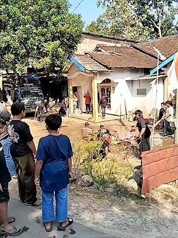 The video shows an outdoor gathering of people near a house. In the foreground, two young boys are facing away from the camera. In the middle ground, several people are gathered around a muddy area where some individuals appear to be lying down. There's a house with a white facade and a brown roof in the background, along with trees and a vehicle. The scene suggests some sort of event or performance is taking place. Further to the right, there is a person holding a red plastic bag. Another part of the video shows a person in traditional attire on the ground, possibly performing, with a decorated truck in the background, equipped with speakers and a canopy. There are other people around the truck, some appearing to be musicians or performers. The overall atmosphere seems to be a lively community gathering.