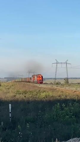 The video shows a train moving along a railway track. The train is red and orange and is emitting a plume of black smoke from its engine. The train is carrying several cargo containers, which are green, orange, and brown. The railway track is surrounded by dry grass and some bushes. In the background, there are several tall electricity pylons with wires stretching across the landscape. The sky is clear and blue, suggesting it is daytime and possibly late afternoon or early evening due to the lighting. The scene appears to be in a rural or undeveloped area.
