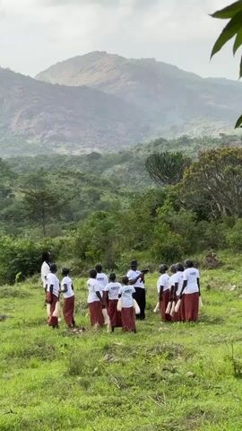 A group of people, mostly women wearing white t-shirts and red skirts, stand in a grassy field, facing inwards towards a person in the center holding a camera. The background consists of lush green hills and mountains under a cloudy sky.
