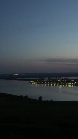 A static shot captures a wide river at dusk, from an elevated position. The sky transitions from a deep blue above to a lighter hue with subtle cloud formations near the horizon. In the distance, a land formation or island is visible, adorned with rows of lights that reflect brightly on the water's surface. The foreground is dominated by a dark, silhouetted hillside with a few sparse trees. Overall, the scene conveys a sense of tranquility and the transition from day to night.