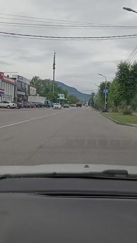 The video shows a view from inside a car driving down a paved road. On the left side of the road, there are buildings, parked cars, and trees. In the distance, there are more cars and a truck on the road, and mountains and trees in the background. The sky is cloudy. The video appears to be shot during the daytime.