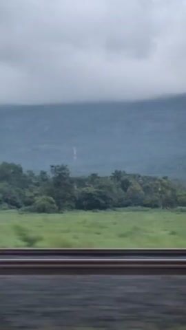 A blurry video taken from a moving train shows a landscape on a cloudy day. In the foreground, train tracks are visible, along with the blurred green fields adjacent to them. In the middle ground, dense green trees form a line, behind which a large mountain range is visible, partially obscured by clouds. There is a very faint indication of a waterfall on the mountainside.
