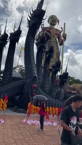 The video begins with a low-angle shot of a large, dark, multi-headed serpent statue with a human-like figure standing on top. A person holding a black umbrella is visible to the left. The scene then pans to the right, showing a row of cave-like structures with various figures inside, including a purple snake-like creature and a mermaid. Children are visible in the foreground. The camera continues to pan, revealing several more figures mounted on animals within the cave-like structures. The shot then shifts to a wider view, showing a large, white, four-faced statue with multiple arms, standing on a pedestal. More cave-like structures and figures are visible in the background. The video then pans to the right again, showing a pathway with people walking. To the left, there is a large grey elephant statue. To the right, there are more cave-like structures with figures, including one that appears to be a dancer. A man wearing a black t-shirt and shorts is walking away from the camera towards the pathway. The sky is cloudy with patches of blue.