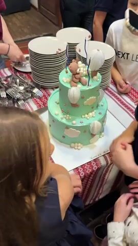 The video shows a two-tiered mint green cake decorated with a teddy bear, hot air balloons, clouds, and pearls. The cake is on a table with a red and white patterned tablecloth. There are stacks of plates, silverware, and people gathered around the cake. In the foreground, a young girl is looking at the cake. To her right, a boy is looking at the cake and smiling. Another young girl is to the left of the cake, also looking at it. The overall scene appears to be a birthday celebration.
