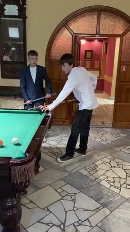 Two young men, one in a white shirt and black pants, the other in a dark suit, are playing billiards. The man in the white shirt is in the foreground, bent over the pool table, preparing to take a shot. The man in the suit is standing to the left, holding a pool cue and watching. The pool table is green with various colored balls scattered on it. The room appears to be a recreation area, with ornate wooden trim, a large mirror on the wall, and arched doorways. The floor is tiled in a mosaic pattern. In the background, another person in a white t-shirt and grey pants is visible.