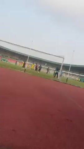 The video shows a soccer practice at a stadium. Players in yellow and dark uniforms are running on the field. The camera is positioned behind the goal, providing a view of the stadium seating in the background. A running track surrounds the soccer field. The sky is overcast. A voice can be heard, though it is not the primary focus of the video.
