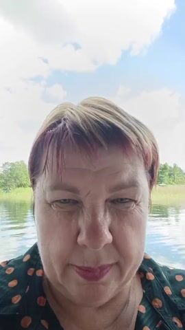 A close-up, head-and-shoulders shot of a middle-aged woman with short, blonde and reddish-brown hair. She is wearing a dark green shirt with orange polka dots. She is looking down towards the camera and smiling slightly. The background is a blurry view of a body of water with trees and a cloudy sky. The video is very short and appears to be a selfie.