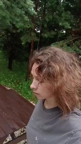 A woman with shoulder-length, wavy, reddish-brown hair is standing outdoors, facing the camera. She is wearing a gray t-shirt. Her expression is neutral. Behind her, there is a wooden picnic table and lush green trees. In the background, a green tent is visible. The ground is covered with grass and some gravel. The overall environment suggests a wooded or park-like setting.