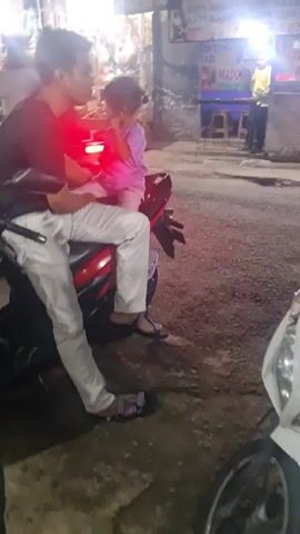 Here is a concise description of the video:

The video shows a man and a young child sitting on a parked motorcycle on a dark street. The man is wearing a black shirt and white pants, while the child wears a purple shirt and pink pants. They appear to be talking to each other. The motorcycle is red and black. In the background, there's a food stall illuminated by bright lights. The surface of the street appears to be paved and a second motorcycle is parked nearby.