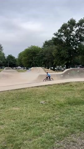 A boy on a bicycle rides through a skatepark. The boy is wearing a blue shirt and black pants. The skatepark has concrete ramps and bowls. There are trees and a building in the background. The sky is cloudy.