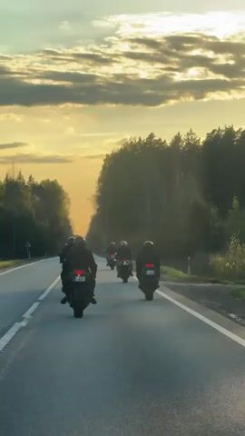 A group of at least six motorcyclists are riding in formation on a gray asphalt road with white lines dividing lanes. They are driving away from the camera. The riders are all wearing helmets and dark clothing. The motorcycles are dark in color and appear to be sport bikes. The road is surrounded by a forest on either side. The sky is a mix of orange, yellow, and gray, indicating either sunrise or sunset, with the sun's rays causing lens flare. A car is seen in the distance ahead of the motorcycles. The video is stable and taken from a lower angle, likely from a vehicle.