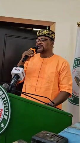 A man in an orange robe and a patterned cap stands at a podium speaking into a microphone. The podium has a green front with a circular logo featuring the words "PUBLIC SERVICE INSTITUTE" and "NIGERIA". A Nigerian flag is visible to the right of the man. A close-up shot reveals names and titles of individuals, including "DR. BERNARD M. DORO" and "ASSOCIATE PROF. BADAMASI M. LAWAL".