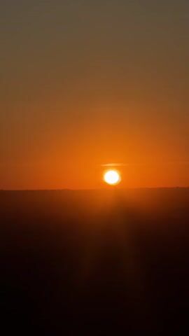 The video shows a sunset. The sun is a bright orange disk near the horizon. The sky is orange and fades to a lighter color at the top. The foreground is dark and blurry, suggesting it is either night or the sun is behind the camera. There are some faint shapes in the distance, possibly trees or buildings. In one shot, a telephone pole is visible on the horizon. The final shot is completely blurry and dark.