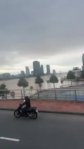 The video depicts a cityscape viewed from a bridge on a cloudy day. A wide river flows between the bridge and the city, with several modern skyscrapers visible in the distance. Motorbikes are driving on the road adjacent to the bridge walkway. A pedestrian walks along the walkway. The bridge has a red brick-lined walkway and a metal safety barrier. There are small trees planted along the walkway. The overall scene suggests an urban environment with a riverfront setting.