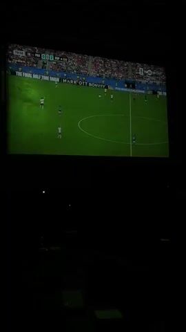 The video displays a projection of a soccer match on a large screen in a dark room. The score on the screen shows "FRA 0:2 ESP". The game is in progress with players on the field. In the foreground, a man wearing a white and blue soccer jersey with French national team insignia is visible, looking intently at the screen with his hand on his chin. Other individuals are partially visible in the background, also appearing to watch the match. The environment is dark, suggesting a viewing party or a cinema-like setting.