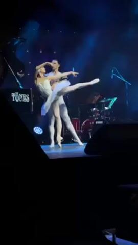 Two ballet dancers, a male and female, are performing on a stage. The female dancer is wearing a white tutu and is supported by the male dancer who is wearing white tights and a black jacket. The female dancer has one leg extended straight up in the air. There are musical instruments in the background, including drums, a piano, and a microphone. There are blue stage lights illuminating the dancers. A speaker partially obscures the view of the stage on the left and right side.