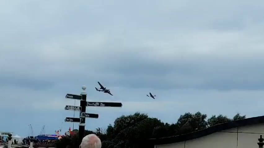 Two twin-engine aircraft fly in formation against a cloudy sky. One plane is larger and closer to the camera, while the other is smaller and further away. Below the planes, a building with a curved facade and the words "THE HERITAGE CENTRE SOUTHPORT" are visible. A street sign and a lamppost are also present in the foreground, along with a person's back. Trees line the background. The planes appear to be performing a flyover or airshow.