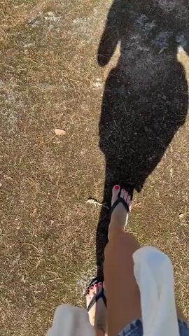 A person's feet are visible in the foreground, wearing flip-flops with painted toenails. A shadow of the person is cast on the ground ahead, indicating they are walking in the direction of the camera. The ground is covered in dry grass and moss. As the video progresses, the perspective shifts to reveal a scenic view of a body of water with sailboats anchored. Lush green trees and rocky terrain surround the water, under a clear blue sky with scattered clouds. The overall scene depicts a person walking outdoors in a natural, coastal environment.