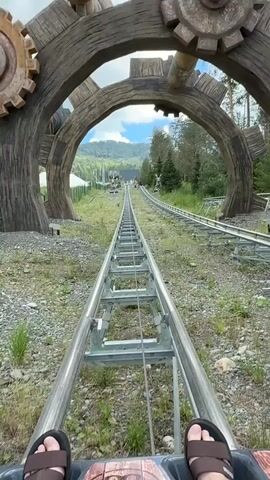 The video begins with a first-person perspective of someone sitting on a roller coaster or alpine slide. The view is focused on the track ahead, which is a metal rail system winding up a gentle slope. On either side of the track, there is overgrown grass and some rocky terrain. In the foreground, the person's sandaled feet are visible. Large, rustic-looking wooden archways with gear-like embellishments frame the scene on the left and right, suggesting a themed attraction. Beyond the immediate track, more of the rail system can be seen extending into the distance, leading up a grassy hillside dotted with trees. In the background, a mountain range with a cloudy sky is visible. The overall environment appears to be an outdoor amusement park or recreational area, possibly in a mountainous or forested region. The footage seems to be captured during the daytime, with natural lighting.