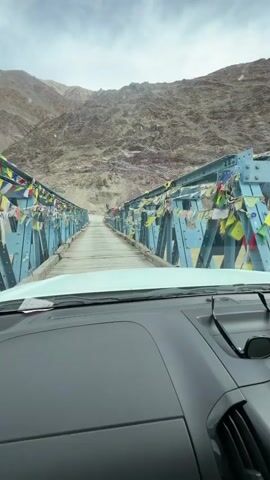 A first-person perspective video shows a vehicle driving across a blue metal bridge. The bridge is adorned with numerous colorful prayer flags on both sides. The bridge spans a muddy, flowing river. Steep, rocky mountains with sparse vegetation rise on either side of the river and frame the view ahead. The sky is cloudy. The interior of the vehicle, including the dashboard and a pair of sunglasses resting on it, is visible in the foreground. The video appears to be taken on a relatively clear day, despite the cloudy sky.