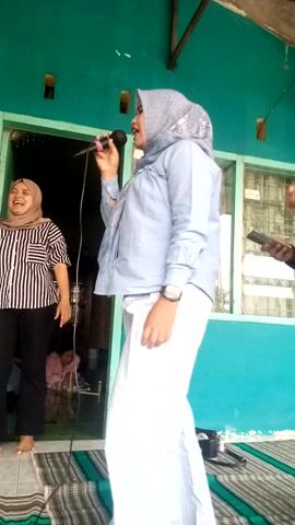 This media file shows an outdoor scene in what appears to be Sukaraja, Indonesia. The dominant color is a vibrant teal green, likely the color of the building walls. In the foreground, two women are the primary subjects. The woman on the left, wearing a light blue hijab and a blue jacket over white striped pants, is holding a microphone and appears to be speaking or singing. She has a wristwatch on her left wrist. Beside her, a woman in a black and white striped top and black pants is also engaged, with her right hand held up and her left hand near her hip. She is wearing a light-colored hijab.

In the background, there are glimpses of another building with green walls and barred windows. A striped rug or mat is visible on the ground, indicating an area possibly set up for an event. Another woman wearing a maroon hijab and black clothing is also visible, holding a mobile phone. The overall atmosphere suggests a lively gathering or performance. The lighting suggests it is daytime, and there are no strong indications of specific weather conditions. The image is oriented vertically.
