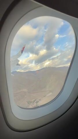 A passenger's view from an airplane window shows a mountainous landscape below. The wing of the plane is visible in the upper left corner of the frame. Clouds are present in the sky, and the sunlight casts a warm glow on the mountains. A river or body of water snakes through the valley below. The terrain appears arid and rugged. The video captures a moment of flight over a remote, natural environment.
