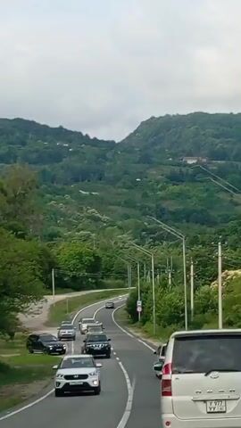 A convoy of vehicles is driving along a winding road through a lush, green mountainous landscape. The road is paved and has white lane markings. Several cars are visible, including a white minivan in the foreground, a white SUV, a black SUV, and several other cars further up the road. Trees and foliage line the road, and houses are visible on the hillsides in the background. The sky is overcast. The video appears to be shot from inside one of the vehicles in the convoy, looking forward. The camera is moving, following the progression of the vehicles.