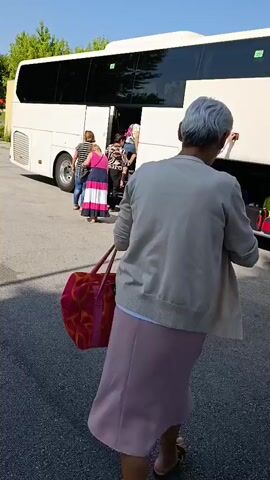 A group of people are boarding a white bus. A woman in a pink dress and a beige cardigan is walking towards the bus with a large pink and orange patterned duffel bag. A man in a white shirt and tie is standing near the luggage compartment of the bus. Another woman in a white shirt and denim shorts is walking past the bus. A woman in a blue dress is standing by the bus door, holding a blue folder. The bus has "ISIDORO DUARTE" written on the side in red letters. The scene appears to be an outdoor setting with trees and a paved area. There is also a white van visible in the background.