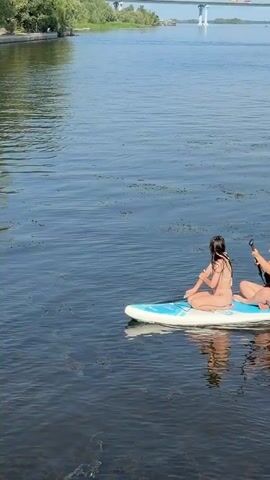 A wide shot shows three young women on a paddleboard on a body of water. The woman on the left is wearing a pink bikini. The woman in the middle is wearing a black bikini and is holding a paddle. The woman on the right is wearing a bikini and is looking off to the side. The water is dark and has some patches of green aquatic vegetation. The sky is light blue. The camera is positioned slightly above and behind the paddleboard.