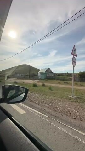 The video is shot from inside a car looking out the passenger side window. The sun is bright and shining in the sky, with some clouds present. A rounded hill is visible in the background, covered in green grass. In the middle ground, there are a few houses with fences. A dirt road runs alongside the car. The car is moving, as indicated by the motion blur and the changing scenery. A side view mirror is visible in the foreground, reflecting the sky and the interior of the car.