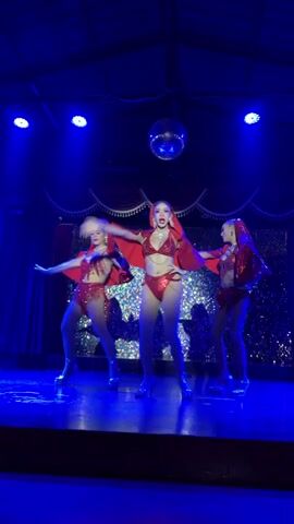 A video captures a stage performance with three individuals dressed in matching red, sparkly outfits. The outfits consist of bikinis with a matching hooded cape or shawl. The individuals are on a stage with blue lighting and a disco ball hanging from the ceiling. The background appears to be a sequined curtain. The individuals are dancing and posing in sync with each other. The central figure is more prominent, with the other two flanking her. The performance seems to be a choreographed dance routine.