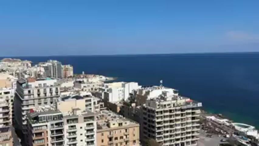 The video shows a high-angle view of a coastal city with several buildings, a green field, and the ocean. The sky is blue with some white clouds. The buildings are mostly white or beige and vary in height. A tall building under construction is visible with a crane on top. The ocean is a deep blue with some areas of turquoise near the rocky shoreline. A road runs along the coast, and a bus is seen driving on it. The video seems to be taken from a balcony or a high vantage point.