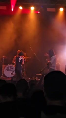 The video shows a band performing on a stage. In the foreground, the audience is visible from behind, with their heads and shoulders filling the bottom of the frame. On stage, a singer in a dark shirt and baseball cap is in the center, holding a microphone. To his right, a drummer is seated behind a drum kit, with a bass drum that says "THE RIGHT PROB...". A person in a white shirt is standing further back on the right side of the stage. The stage is lit with red and yellow lights, and there is a significant amount of stage smoke. The overall atmosphere appears to be a live concert.
