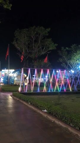 The scene is an outdoor nightscape. A walkway leads to a grassy area where decorative triangular structures are illuminated with multicolored lights. Behind the structures, three red flags are mounted on poles. Trees and other light installations are also visible in the background.