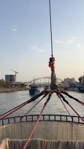 The video begins with a close-up shot of a fishing net and ropes on a boat. The perspective shifts to show a river with several boats, including a blue fishing boat in the distance. In the background, a bridge and buildings are visible, along with a construction crane. The sky is clear and blue, with a few clouds. The video is filmed from a moving boat, suggesting a journey on the river. The overall scene depicts a harbor or riverfront area, likely in a coastal city.