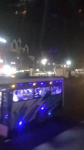 At night, a parking lot is filled with rows of parked motorcycles. Streetlights illuminate the area, and the windows of a nearby building with a gabled roof are visible. A bus pulls away from the curb, its interior lights revealing passengers inside. A few people are walking around the parking lot. The ground is dark.