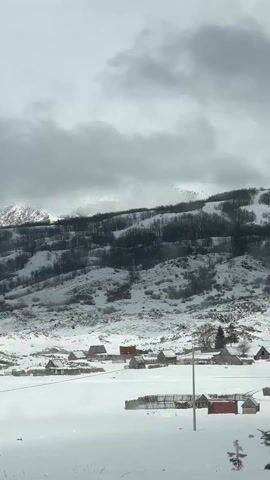 The video shows a snow-covered village landscape under a cloudy, overcast sky. Several small, wooden houses with red roofs dot the foreground, surrounded by a thick blanket of snow. A rudimentary wooden fence encloses a few of the properties. In the background, a snow-covered mountain range is visible, with dark trees sparsely covering its slopes. The overall atmosphere is cold and isolated, accentuated by the muted colors and heavy cloud cover.