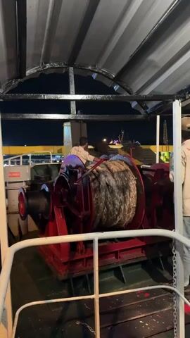 The media file depicts a scene on a ferry at night, likely in Ketapang, East Java. The primary focus is a large, red winch with a thick, coiled rope. A person in a light-colored shirt and jeans stands behind the winch, seemingly interacting with it. Another person, partially visible on the right side of the frame, wears a light-colored jacket and jeans.

The setting appears to be the deck of the ferry, under a covered structure with a corrugated metal roof supported by dark beams. The background reveals the dark night sky and the illuminated structures of another vessel or the port in the distance. Light poles and a tower-like structure are visible, suggesting a port environment.

The activity appears to be the operation or preparation for operating the winch, possibly for docking or departing the ferry. The presence of the winch and rope strongly indicates maritime activity. The lighting suggests it is nighttime. There are no overt indications of weather conditions, but the covered structure might imply protection from elements. The mood is functional and task-oriented, with no discernible emotional expression from the visible individuals.

A red sign with white text, partially obscured, reads "BLOWER" on a white object to the left of the winch. The overall scene is utilitarian, typical of a working ferry or ship deck.