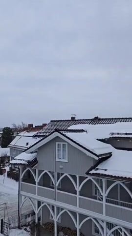 The video shows a snowy residential neighborhood on an overcast day. The camera pans across various houses, most of which are white or gray, and their roofs are covered with snow. A church steeple is visible in the distance. The streets and yards are also covered in snow. There are bare trees scattered throughout the neighborhood.