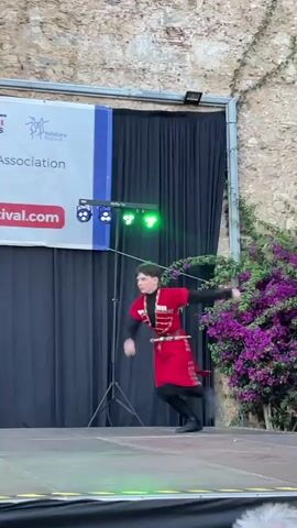 A young male dancer in a red traditional costume performs on a stage. The costume includes a red tunic with gold trim, black leggings, and black boots. He is in mid-air, leaping with his arms extended. In the background, a banner for the "Folk Festival Association" is visible. Several other performers in similar red costumes are standing on the stage behind the main dancer. The backdrop is a stone wall adorned with purple bougainvillea. The stage is illuminated by stage lighting. Spectators can be seen in the foreground, holding up phones to record the performance.