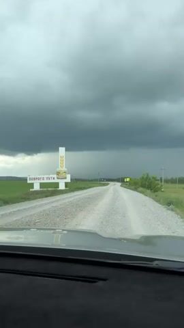 A view from inside a moving vehicle on a dirt road shows a dark grey sky, green fields, and a white welcome sign with "ДОБРОГО ПУТИ" (Bon Voyage) written on it. The sign is adorned with an image of golden wheat ears. The road ahead curves slightly to the right, and what appears to be a heavy rain shower can be seen in the distance, falling from the dark clouds.