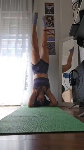 A woman is doing a headstand on a green yoga mat. Her feet are up against a white wall, and she is wearing a matching athletic outfit of gray shorts and a sports bra. She has socks on. Behind her, on the wall, are three framed posters: a KISS poster, a Jimi Hendrix poster, and a Led Zeppelin poster. To the right of the woman, there is a mirror, a desk, a television, and a pair of headphones. The floor is made of wood.