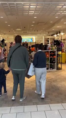 The video begins inside a brightly lit retail store filled with stuffed animals. A person from behind, wearing a green tracksuit and a black crossbody bag, holds the hand of a small child dressed in a dark blue outfit. To their right, a woman wearing a blue sweater and striped pants, holding a white shopping bag, is seen from behind. The store's shelves are stocked with various plush toys, including Halloween-themed items. The video then pans to the right, showing more of the store's merchandise and then moves to show the exterior of the store, revealing a mall corridor. The corridor is tiled and has several closed storefronts. A mannequin in a yellow shirt and jeans stands in a display case, and a poster is visible on a pillar. The camera continues to pan, showing more of the empty mall.