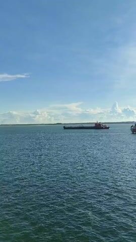 The video captures a wide shot of a body of water under a partly cloudy blue sky. Two large cargo ships are visible in the distance. The ship on the left is dark-colored with a red superstructure, while the ship on the right is predominantly white with an orange hull. The water is choppy, reflecting the sunlight. The horizon is flat, suggesting an open sea or large bay. The camera appears to be panning slowly to the right. No people are visible, and no specific actions are clearly discernible beyond the ships moving slowly on the water. The overall scene depicts a maritime environment on a clear day.