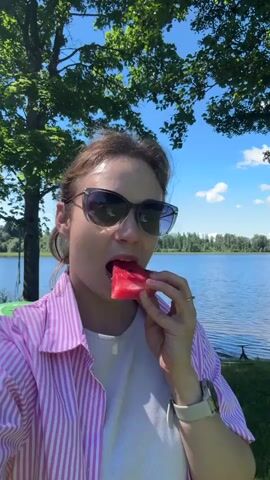 A young woman with sunglasses and a pink and white striped shirt is eating a slice of watermelon outdoors. She is standing in front of a body of water with trees and a blue sky with clouds in the background. The woman is holding the watermelon in her right hand and taking a bite. The video is a selfie, as evidenced by the woman holding the camera herself.